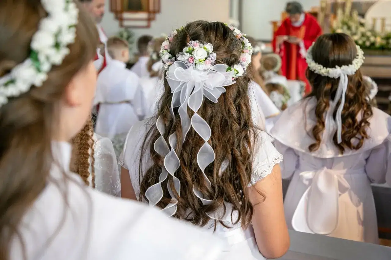 Dziewczynki w białych strojach i wiankach czekają na komunię świętą. Włosy ozdobione wstążkami.