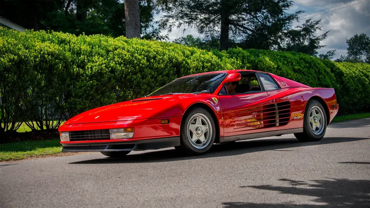 Czerwone Ferrari Testarossa z lat 80. błyszczy na tle zielonego żywopłotu.