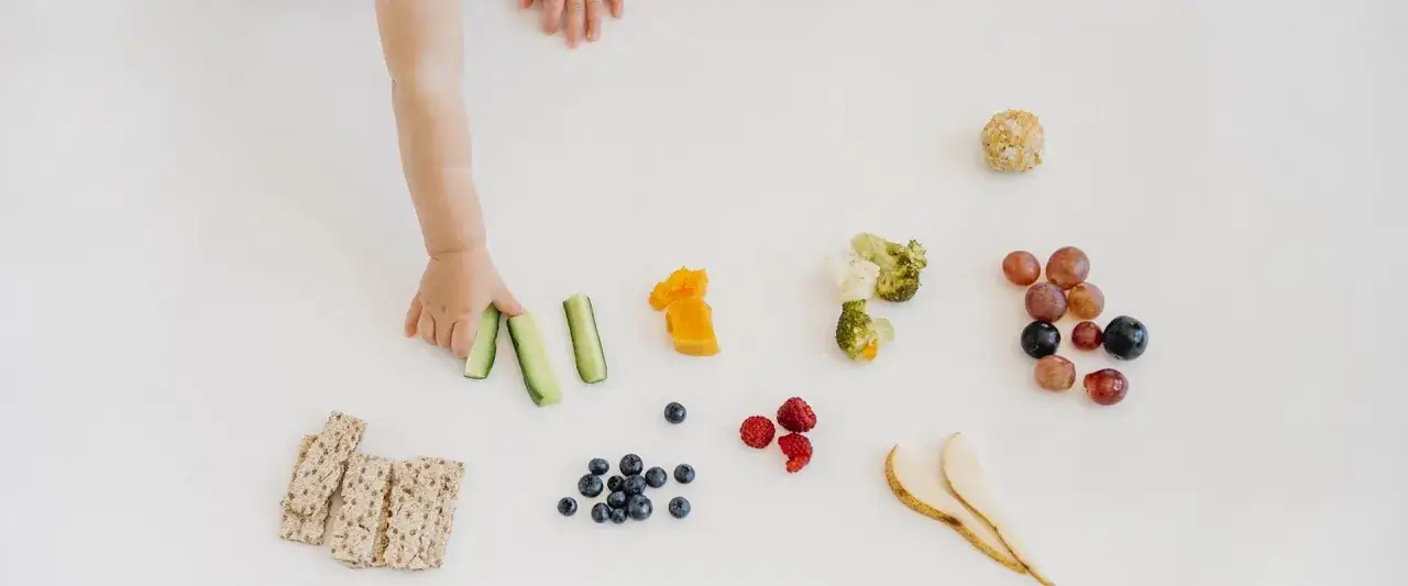 Mano di bimbo esplora cibo per svezzamento: cetrioli, zucca, broccoli, mirtilli, lamponi, uva, pere e cracker. Tagli sicuri svezzamento.