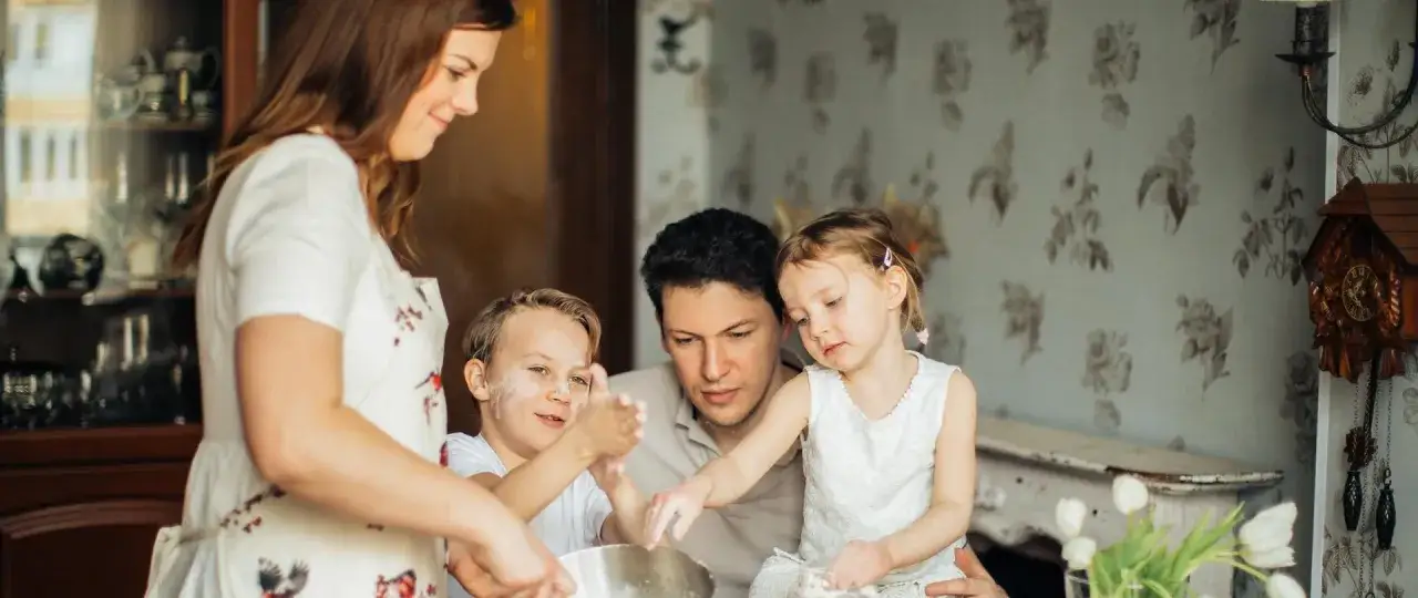 Rodzina aktywnie w domu: mama, tata i dwójka dzieci piecze ciasto. Chłopiec ma mąkę na twarzy, dziewczynka patrzy z uwagą.