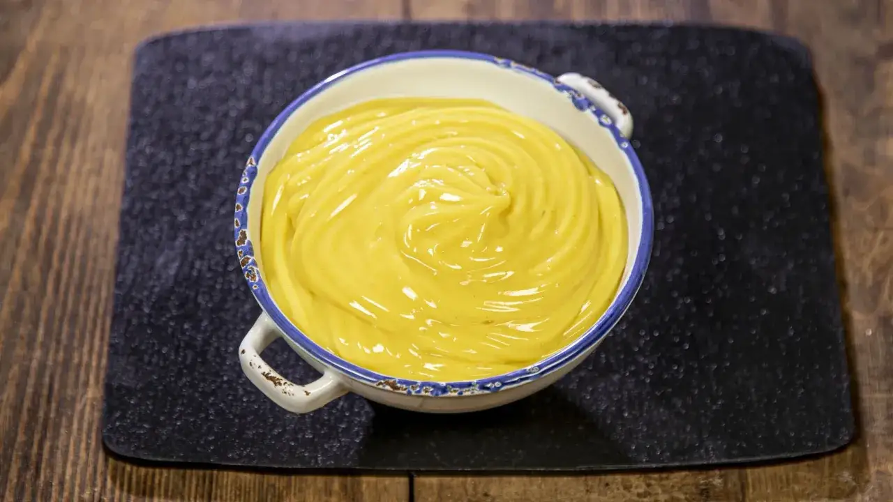 Una ciotola di crema pasticcera vegana, liscia e gialla, con un bordo blu e bianco, su un tappetino nero.