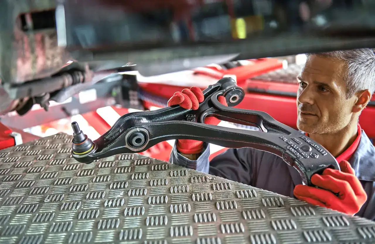 Mechanik w czerwonych rękawiczkach prezentuje nowy wahacz, kluczowy element zawieszenia, który zapewni stabilizatorowi pojazdu lepszą przyczepność.