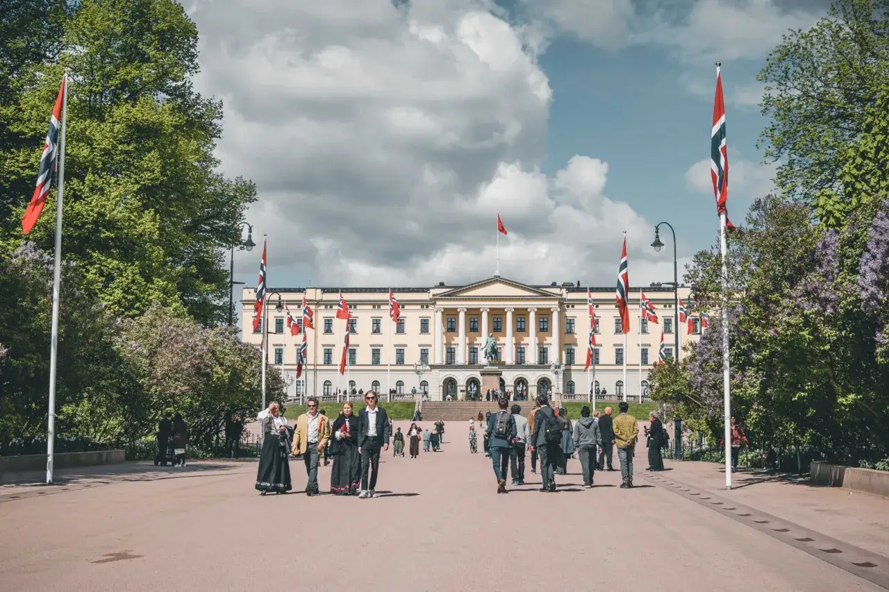 Przed Pałacem Kr&oacute;lewskim w Oslo, z norweskimi flagami, spacerują ludzie. To miejsce, gdzie można poczuć atmosferę norweskiej rodziny kr&oacute;lewskiej.