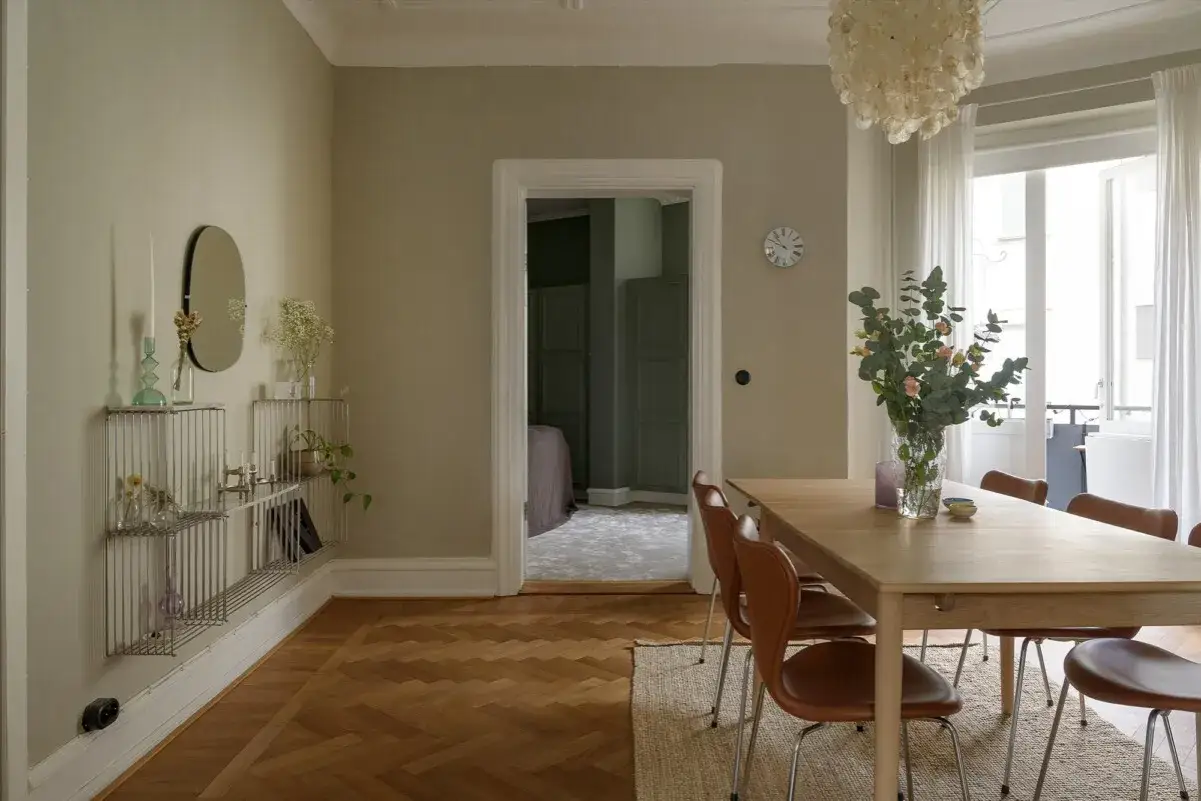 Comedor con mesa de madera, sillas marrones y pared color crema. Un jarr&oacute;n con flores decora la mesa.