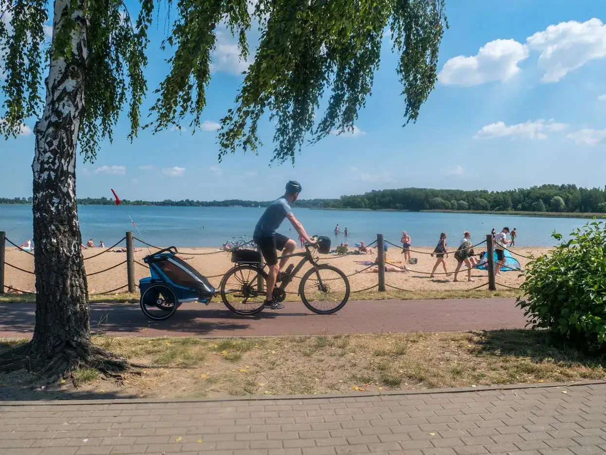 Rowerzysta z przyczepką przemierza malownicze trasy rowerowe Śląska, obok plaży z wypoczywającymi ludźmi.