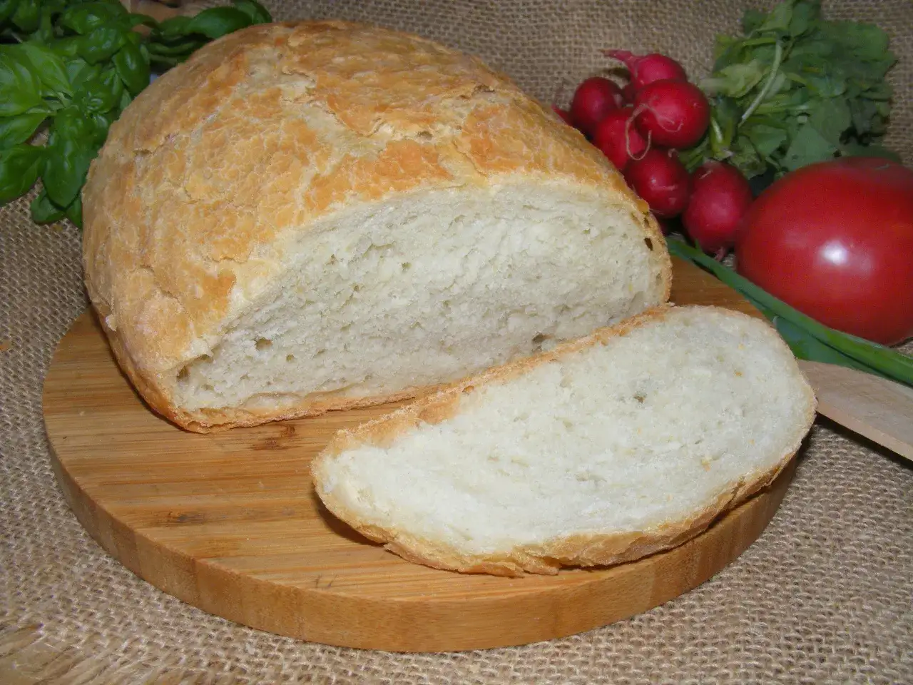 Pyszny, domowy chleb z garnka, pokrojony na desce, z pomidorem i rzodkiewkami w tle.