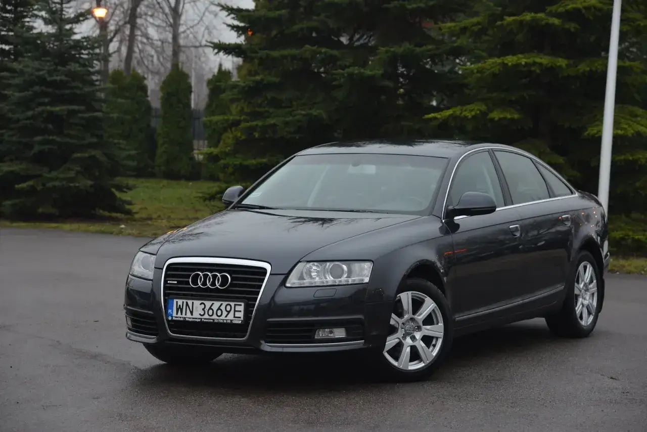 Ciemnoszary Audi C6 na mokrej drodze, otoczony zielenią drzew.