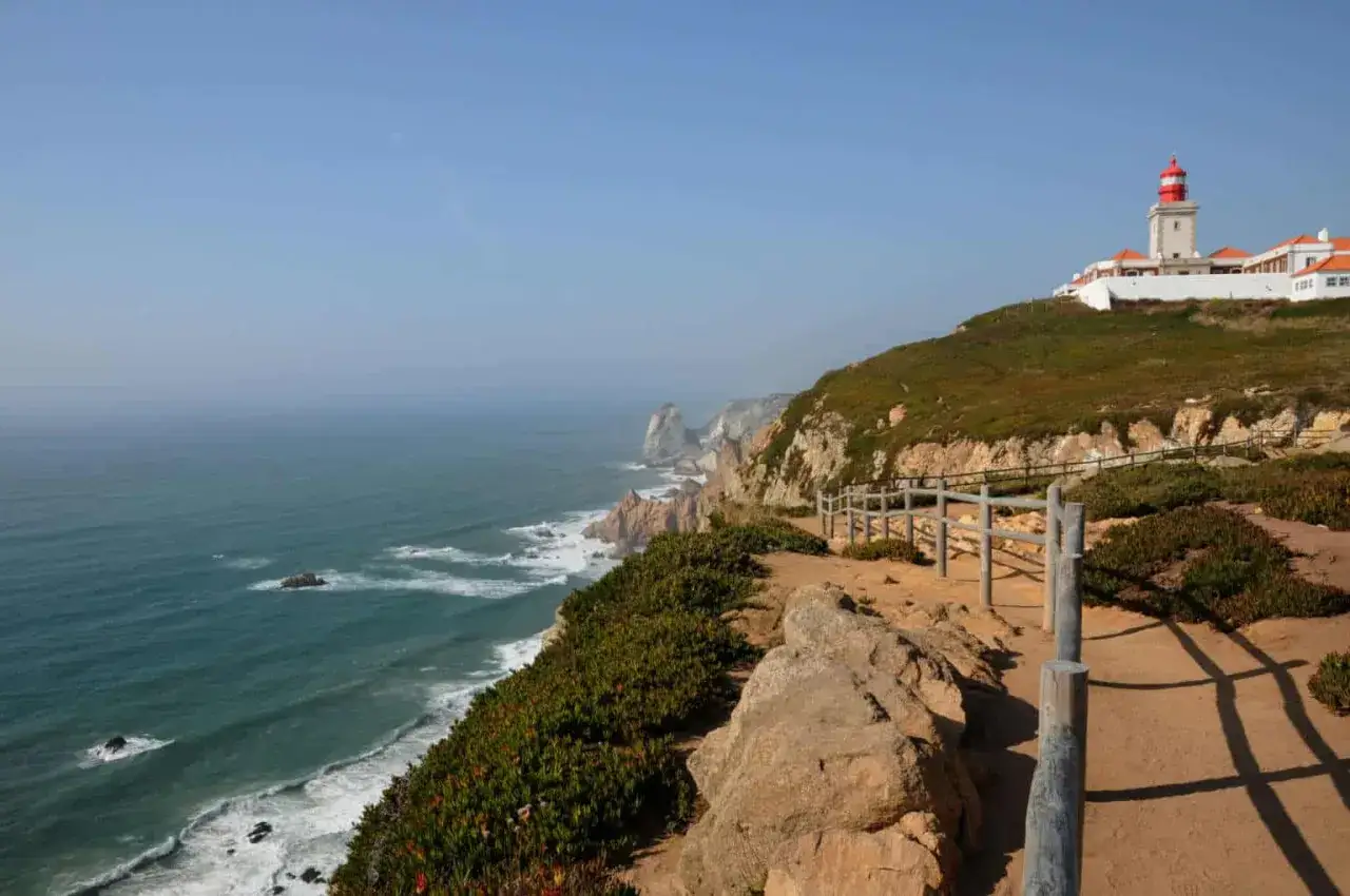 Widok na ocean i klify z latarnią morską na Cabo da Roca. Ścieżka z drewnianym płotkiem prowadzi wzdłuż wybrzeża.