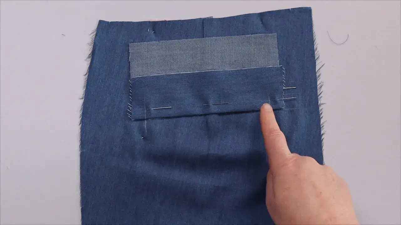 A finger points to a partially constructed back welt pocket on dark blue fabric. Pins hold the pocket in place.