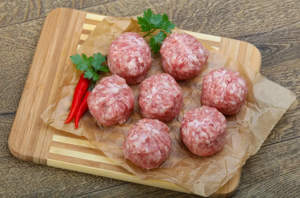 Siedem okrągłych kotlecików mielonych bez bułki, ułożonych na papierze do pieczenia, obok dwóch czerwonych papryczek chili i gałązki pietruszki.