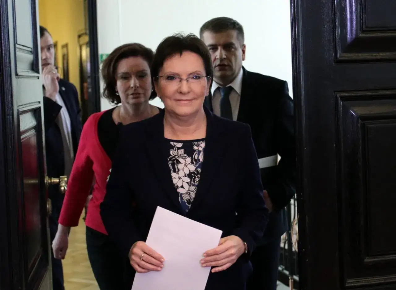 Ewa Kopacz premier, w towarzystwie innych osób, wychodzi zza drzwi, trzymając dokumenty.