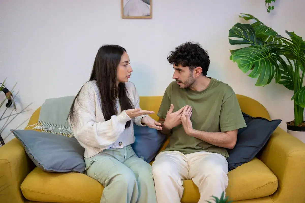 Ein Paar sitzt auf einem gelben Sofa und diskutiert angeregt. Ihre K&ouml;rpersprache deutet auf **partnerschaftliche Probleme** hin, w&auml;hrend sie versuchen, eine L&ouml;sung zu finden.