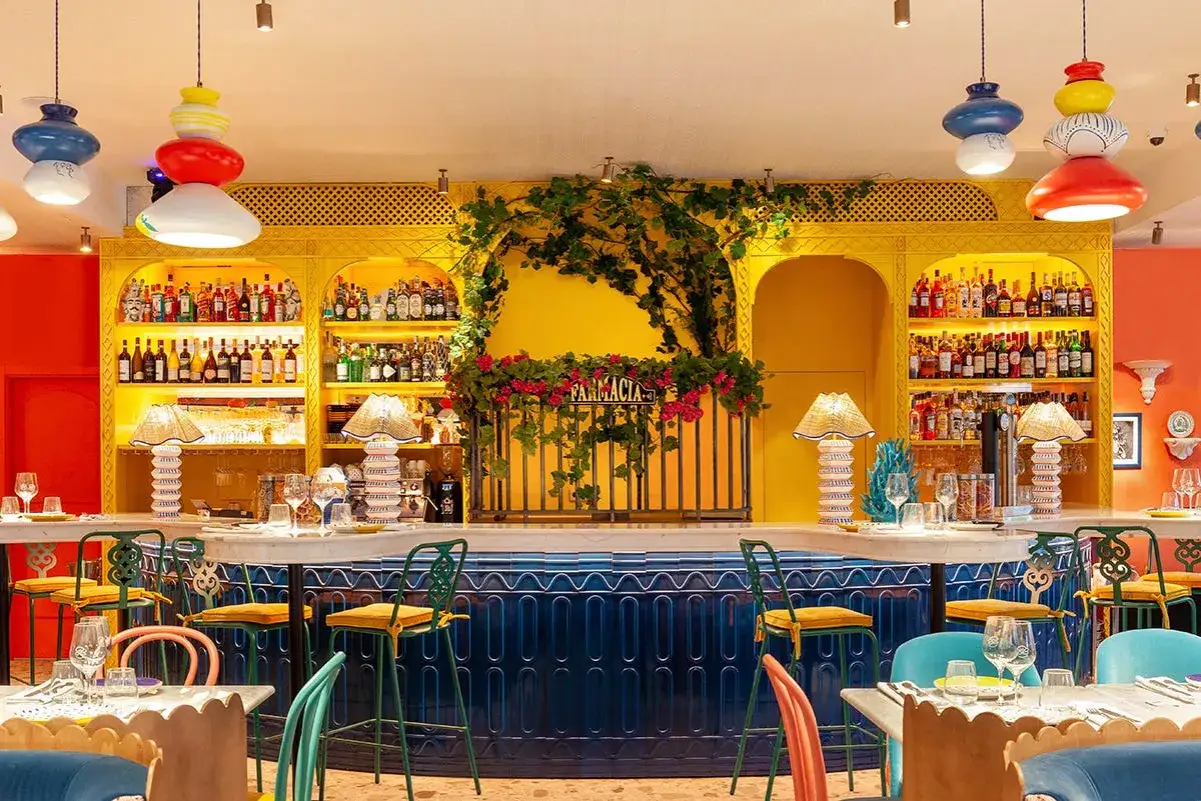 Bar vibrante con estanter&iacute;as amarillas llenas de botellas, l&aacute;mparas de mimbre y sillas coloridas. Un lugar perfecto para disfrutar de una beata pasta.