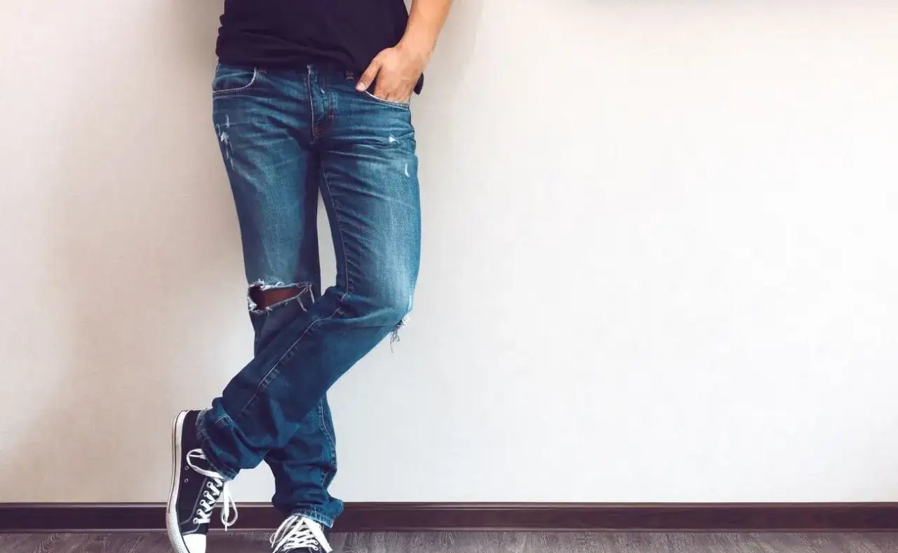 Mężczyzna w modnych jeansach męskich z dziurami na kolanie, czarnych trampkach i czarnej koszulce.
