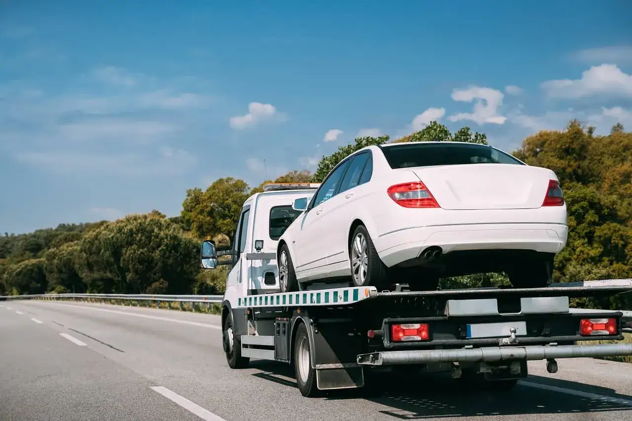 Biały samochód jest transportowany na lawecie. To pokazuje, czy można holować samochód w takiej sytuacji.