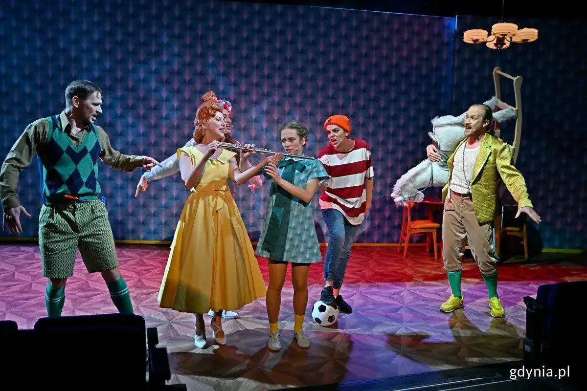 Scena teatralna pełna barw i ruchu. Kobieta w żółtej sukni gra na flecie, towarzyszy jej mężczyzna w kamizelce. W tle widać postacie w strojach przypominających bajkowe, tworzące radosne scherzo.