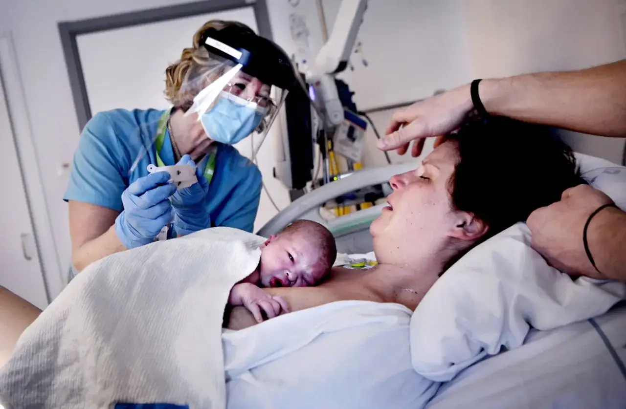 Nyfödd bebis på bröstet hos en nyförlöst mamma. En sköterska i skyddsutrustning håller i ett instrument. En pappa tröstar. GBS gravid.