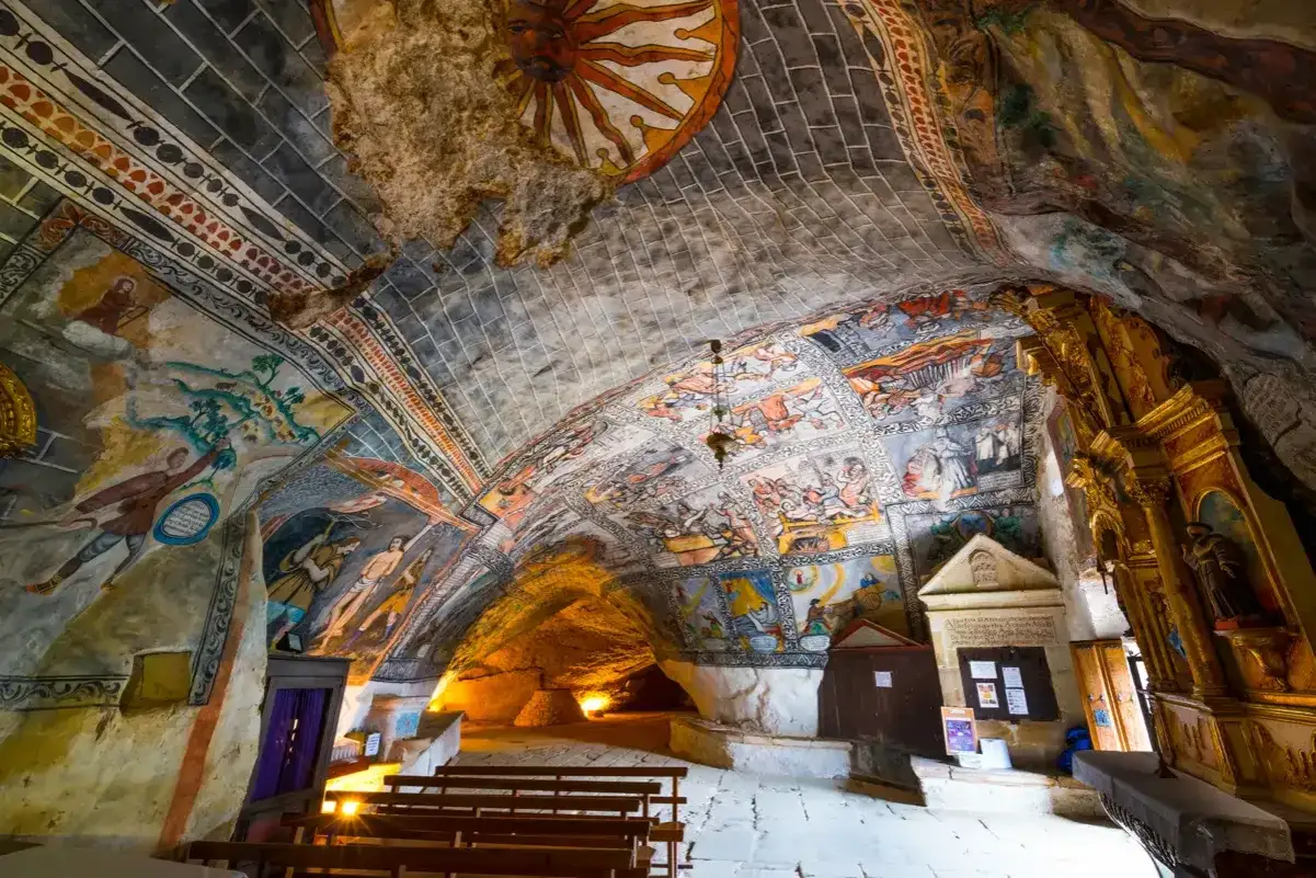Interior de una ermita con pinturas murales que narran historias bíblicas, un sol en el techo y un ojo guareña que vigila desde la roca.
