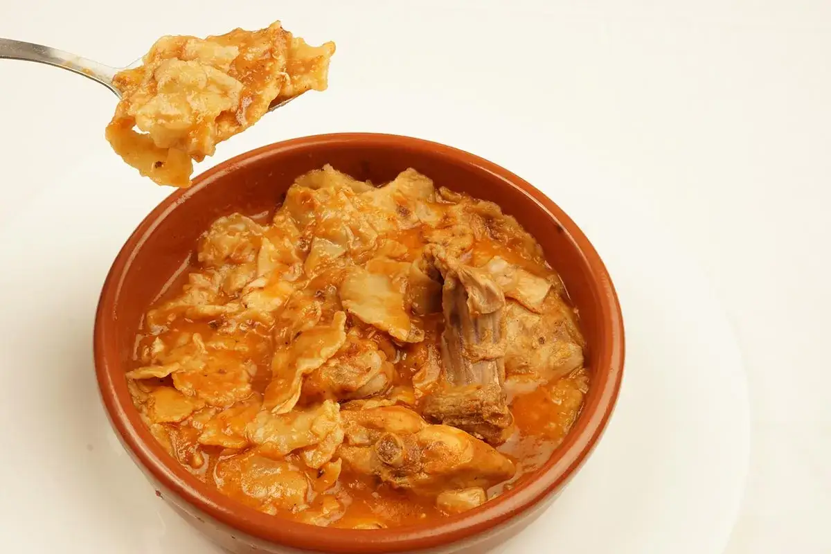 Un plato humeante de comida típica de Albacete, con trozos de carne y pasta en una salsa rojiza.