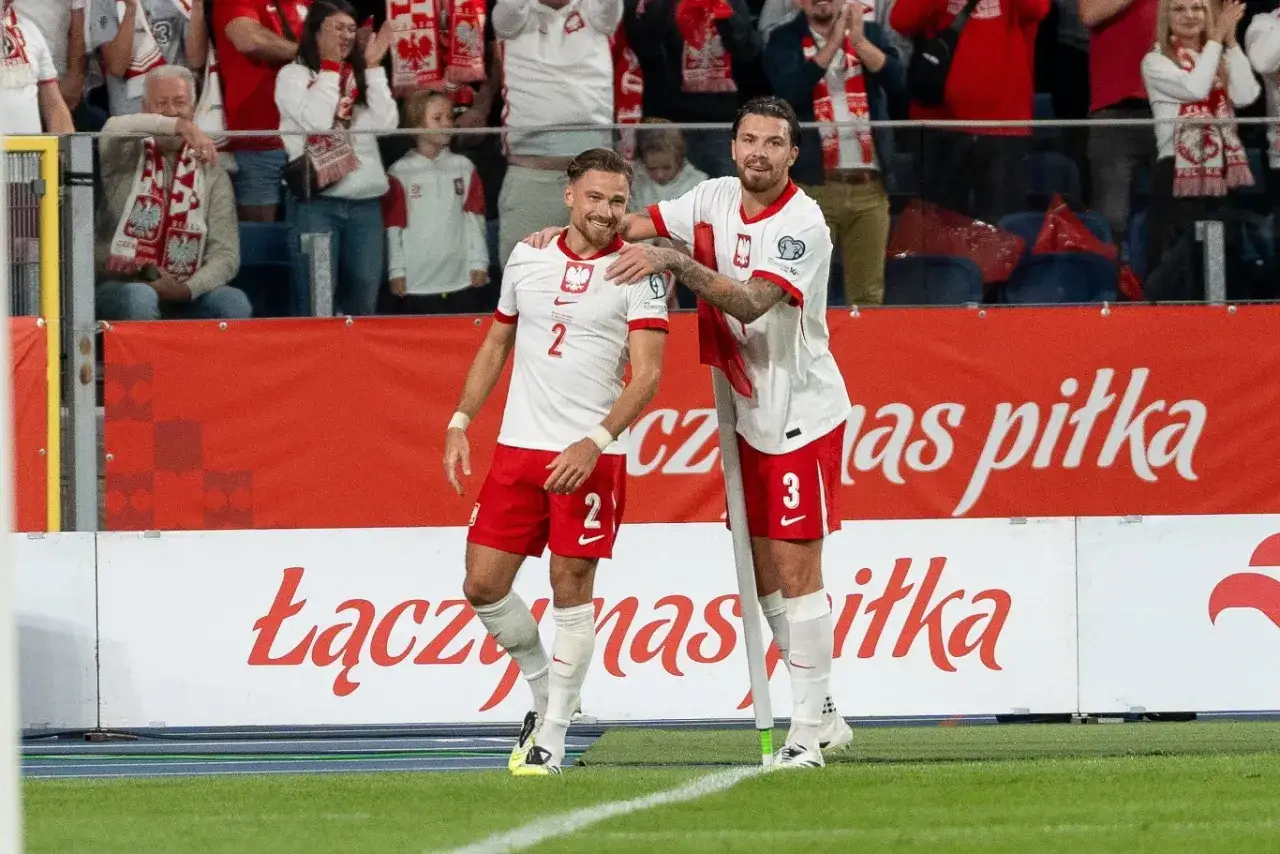 Dwóch piłkarzy w biało-czerwonych strojach cieszy się na boisku. Jeden z nich, z numerem 2, ma rękę na ramieniu kolegi. Obaj chcą grać dla Polski, czuć dumę i wsparcie kibiców.