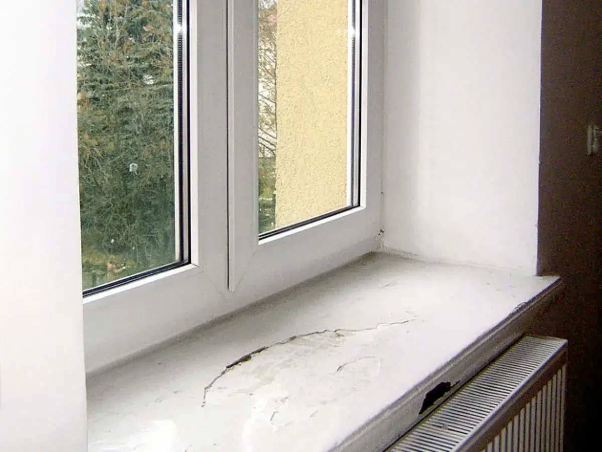 Uszkodzony parapet wewnętrzny okna, wymaga wymiany. Widoczny pęknięty biały parapet i grzejnik.