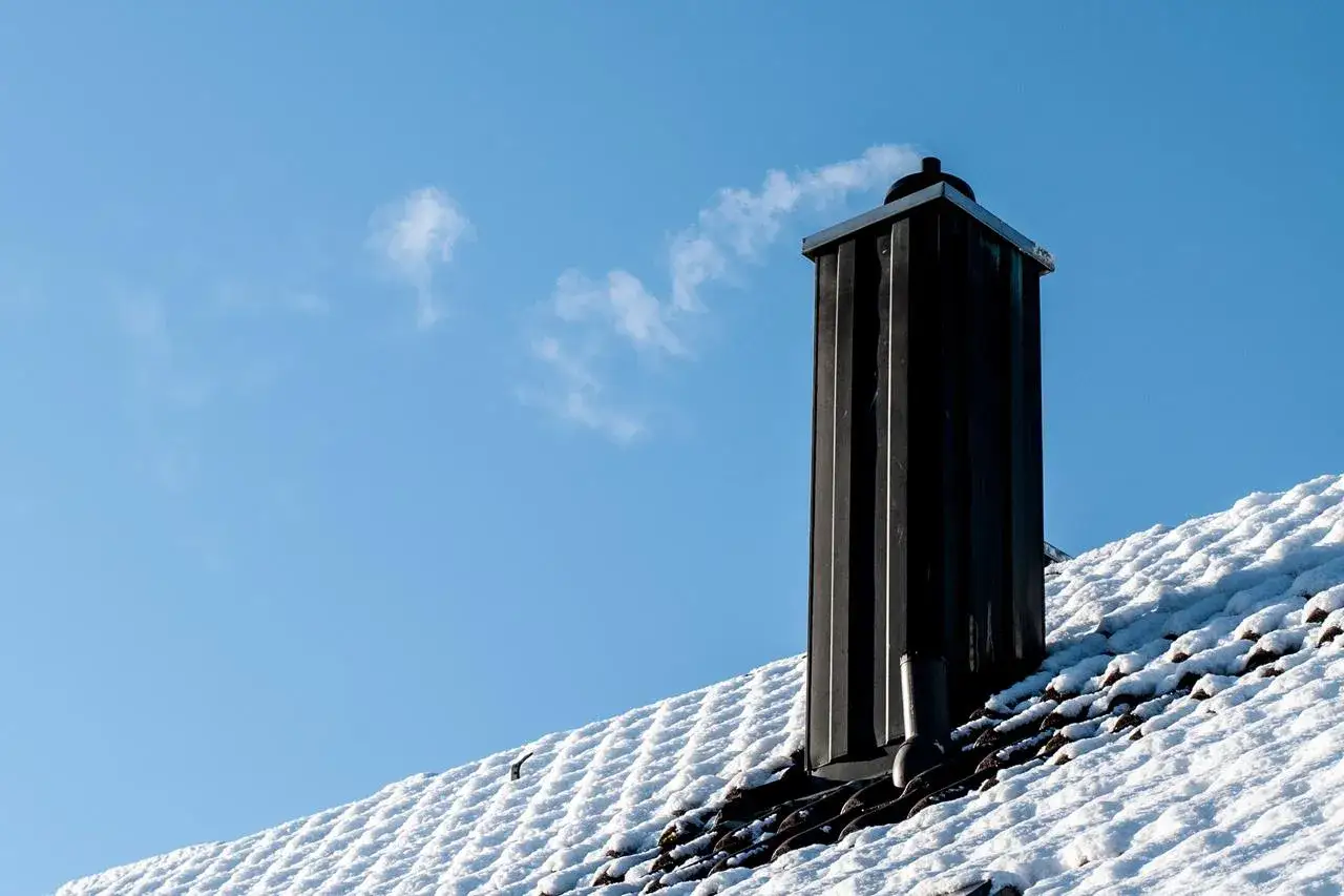 En svart skorsten avger r&ouml;k mot en klarbl&aring; himmel. Taket &auml;r t&auml;ckt av sn&ouml;, vilket ger en k&auml;nsla av kallt v&auml;der.