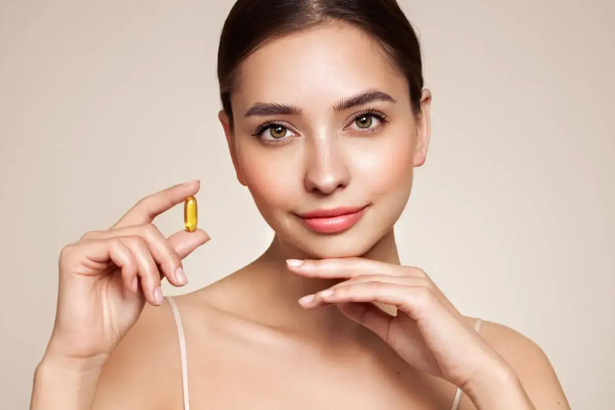 Mujer sonriente sostiene una cápsula dorada, un secreto para la piel radiante. Vitamina E para la cara, un toque de juventud.