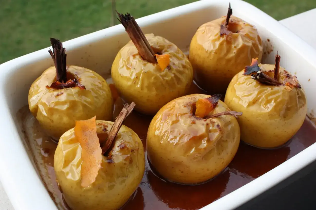 Seis manzanas asadas doradas, con canela y piel de naranja, reposan en una bandeja blanca con un sirope oscuro.
