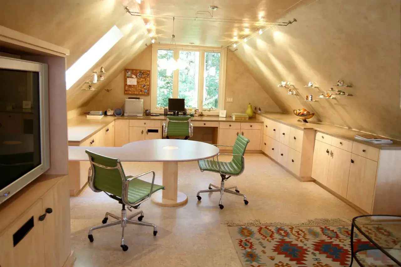 Acogedor despacho en buhardilla con muebles de madera clara, sillas verdes y una mesa ovalada. Luz natural entra por la ventana.