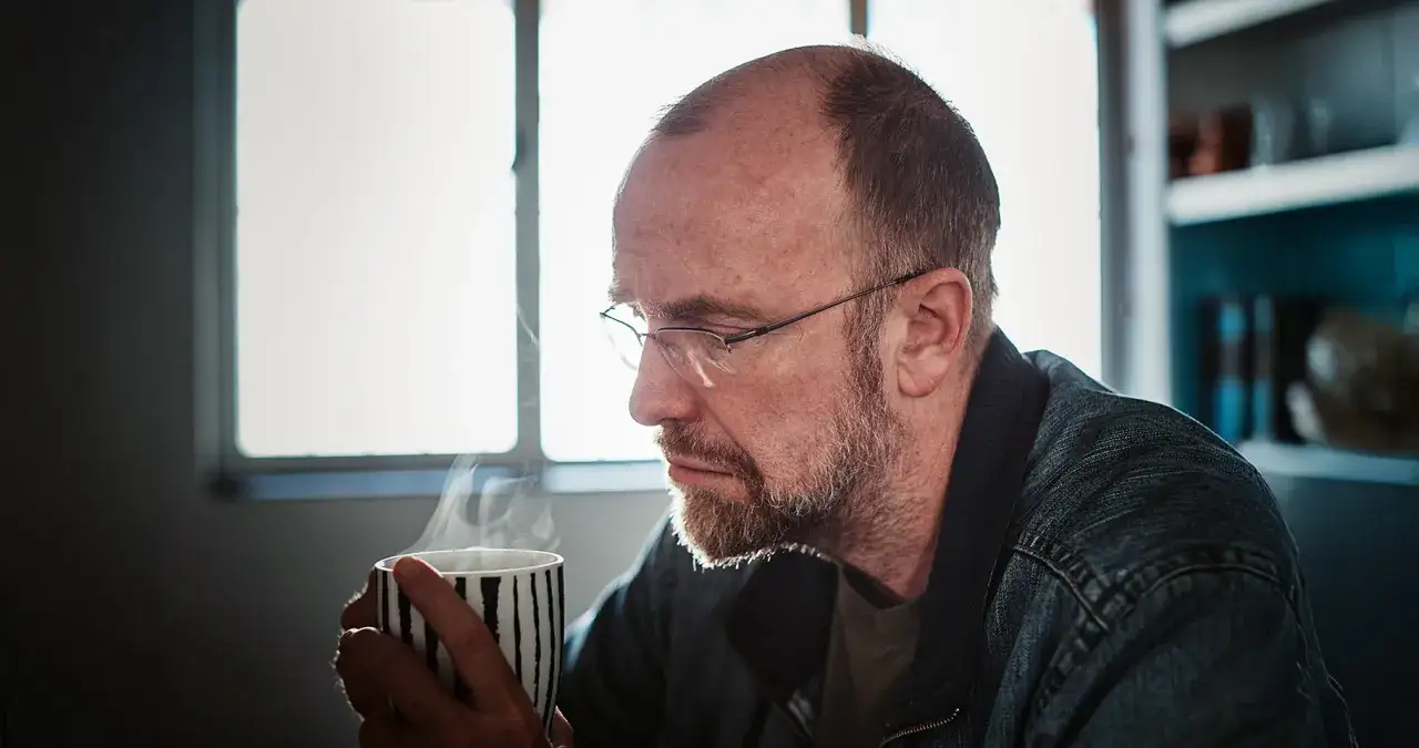 Ein nachdenklicher Mann mit Brille hält eine dampfende Tasse. Er wirkt wie ein Mann in der Midlife-Crisis, der über das Leben sinniert.