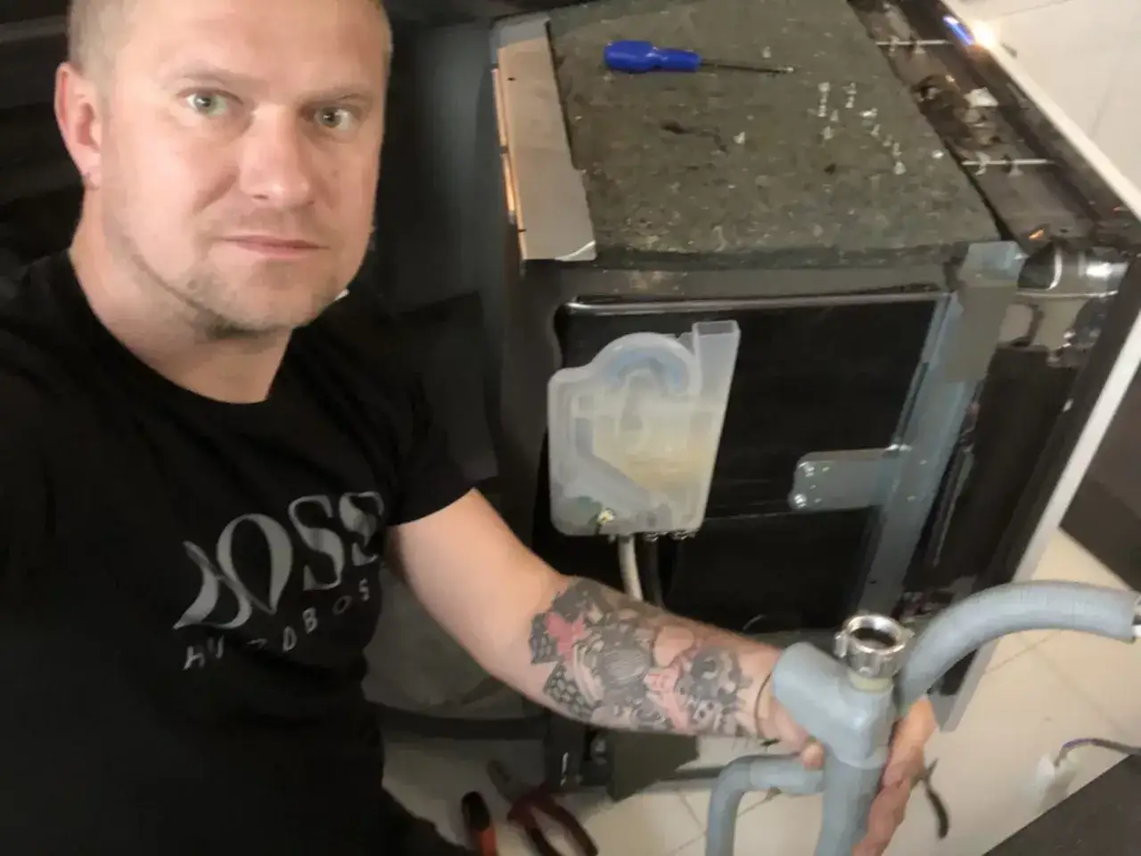 Mężczyzna naprawia zmywarkę Whirlpool z błędem E4. Widoczny jest zbiornik na wodę i wąż odpływowy.