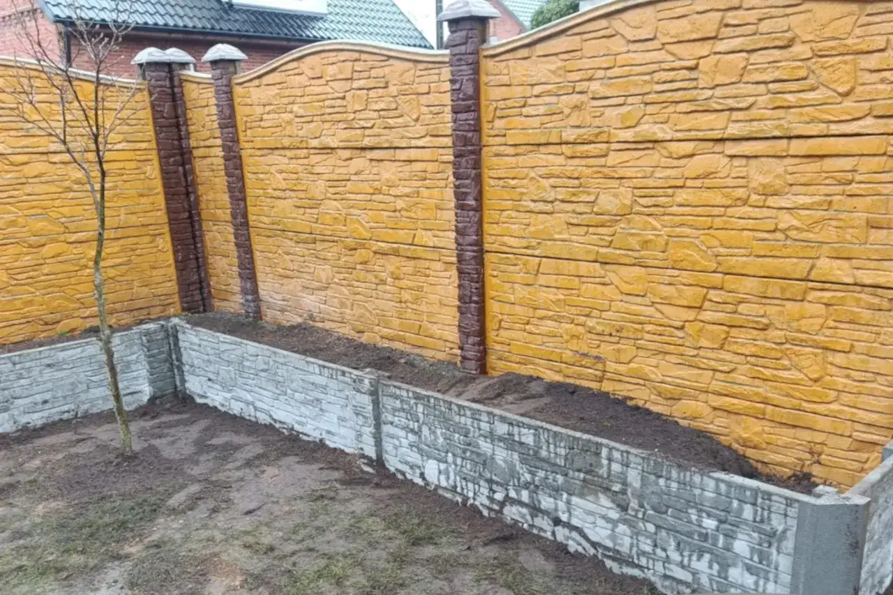 Żółte ogrodzenie betonowe z brązowymi słupkami. Niska betonowa rabata przed nim. Zastanawiasz się, na jaki kolor pomalować ogrodzenie betonowe?