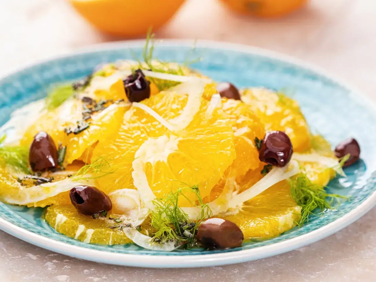 Erfrischender Salat mit saftigen Orangenscheiben, garniert mit dunklen Oliven und Fenchelgrün auf einem blauen Teller.