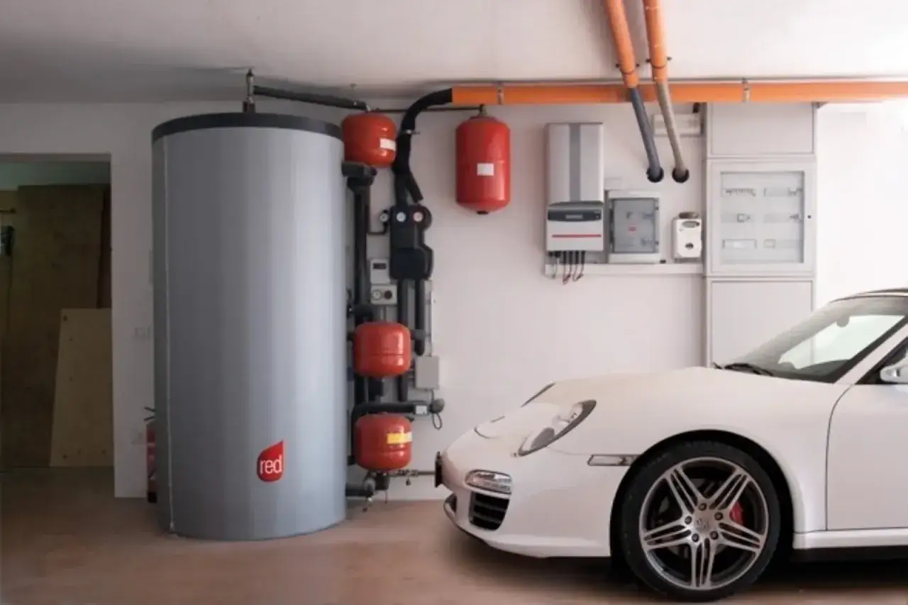 Impianto di riscaldamento con grande puffer per caldaia a pellet, serbatoi rossi e quadro elettrico, accanto a una Porsche bianca.