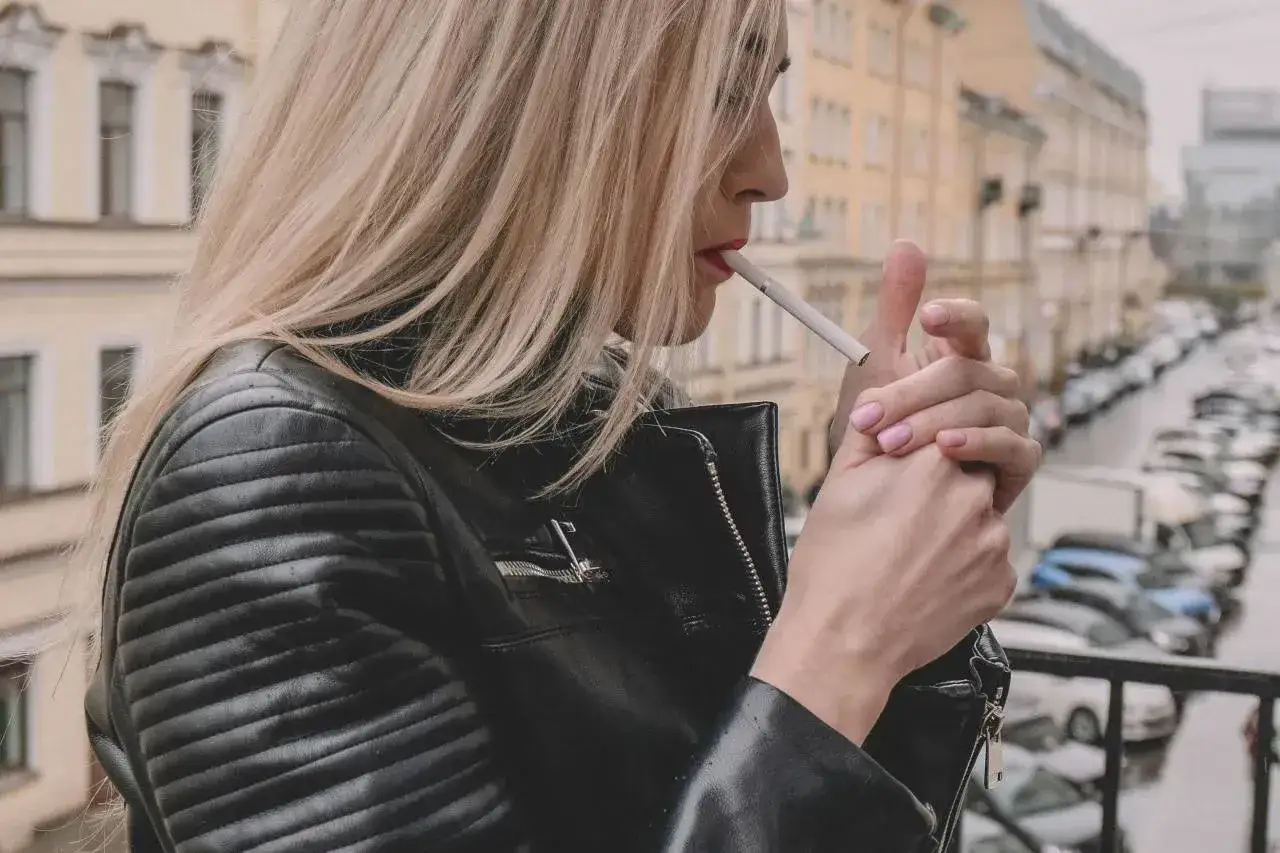 Kobieta w skórzanej kurtce zapala papierosa na balkonie, zastanawiając się, czy można palić na balkonie.