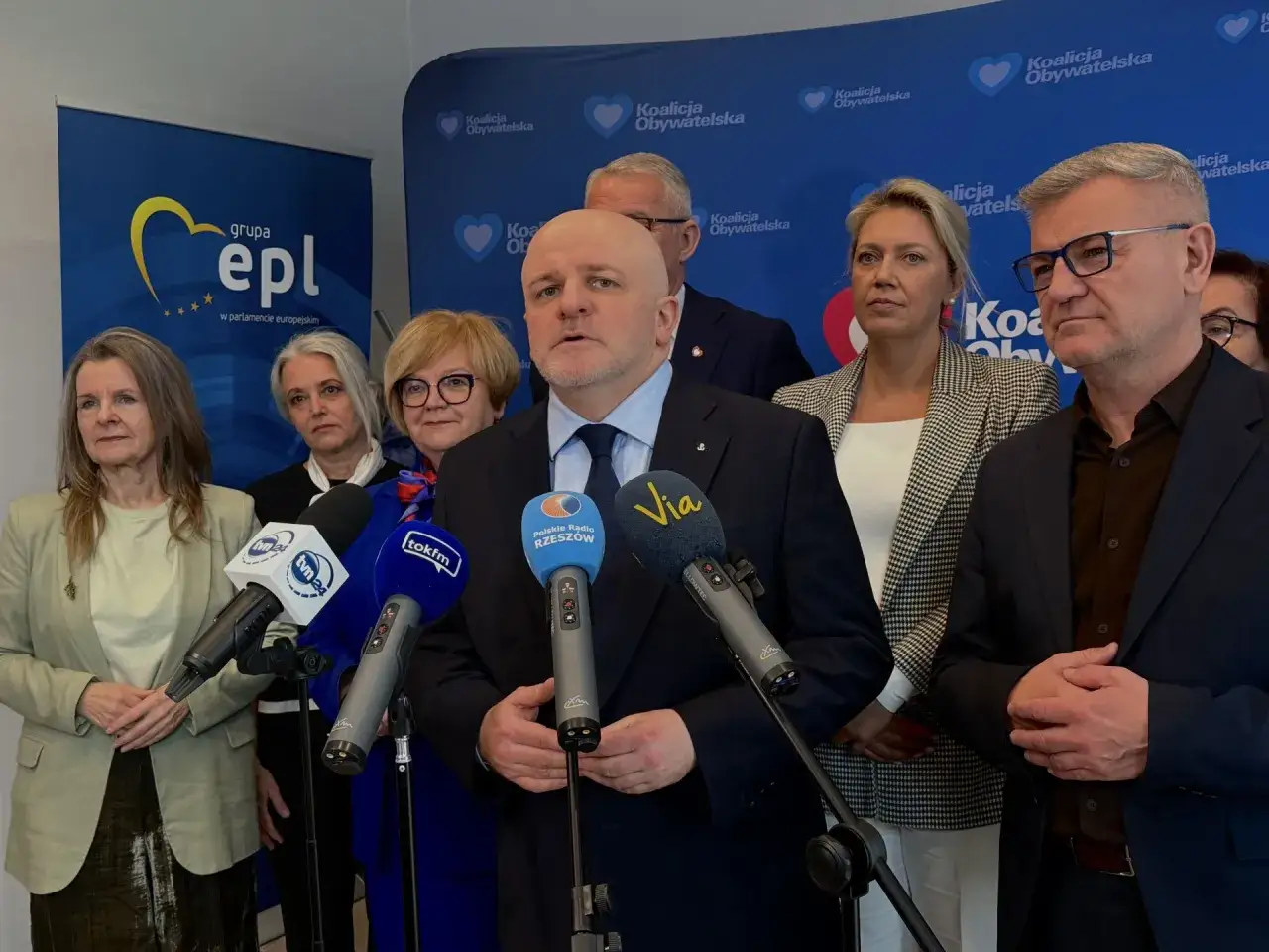 Konferencja prasowa Koalicji Obywatelskiej. Mężczyzna w garniturze przemawia do mikrofonów, obok stoją kobiety i mężczyźni.