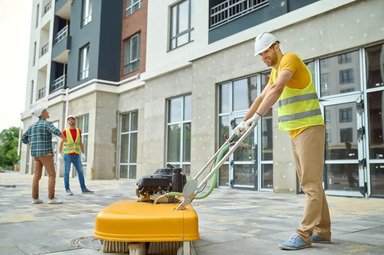 Operai impegnati nella pulizia post cantiere con una spazzatrice meccanica. L'edificio moderno sullo sfondo è quasi completato.