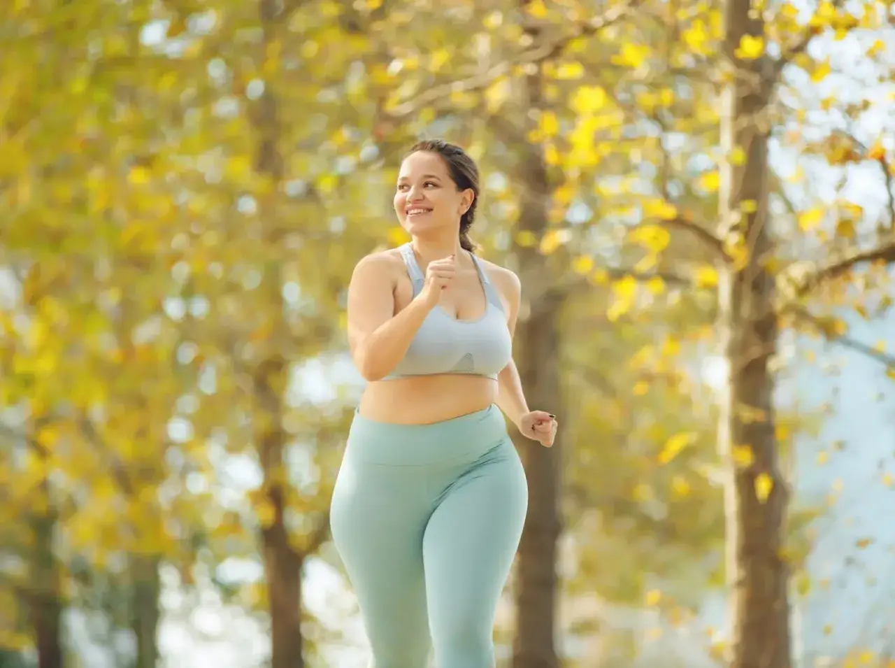 L&auml;chelnde Frau joggt im Herbstwald. Sie tr&auml;gt Sportkleidung und genie&szlig;t das Laufen, um abzunehmen.