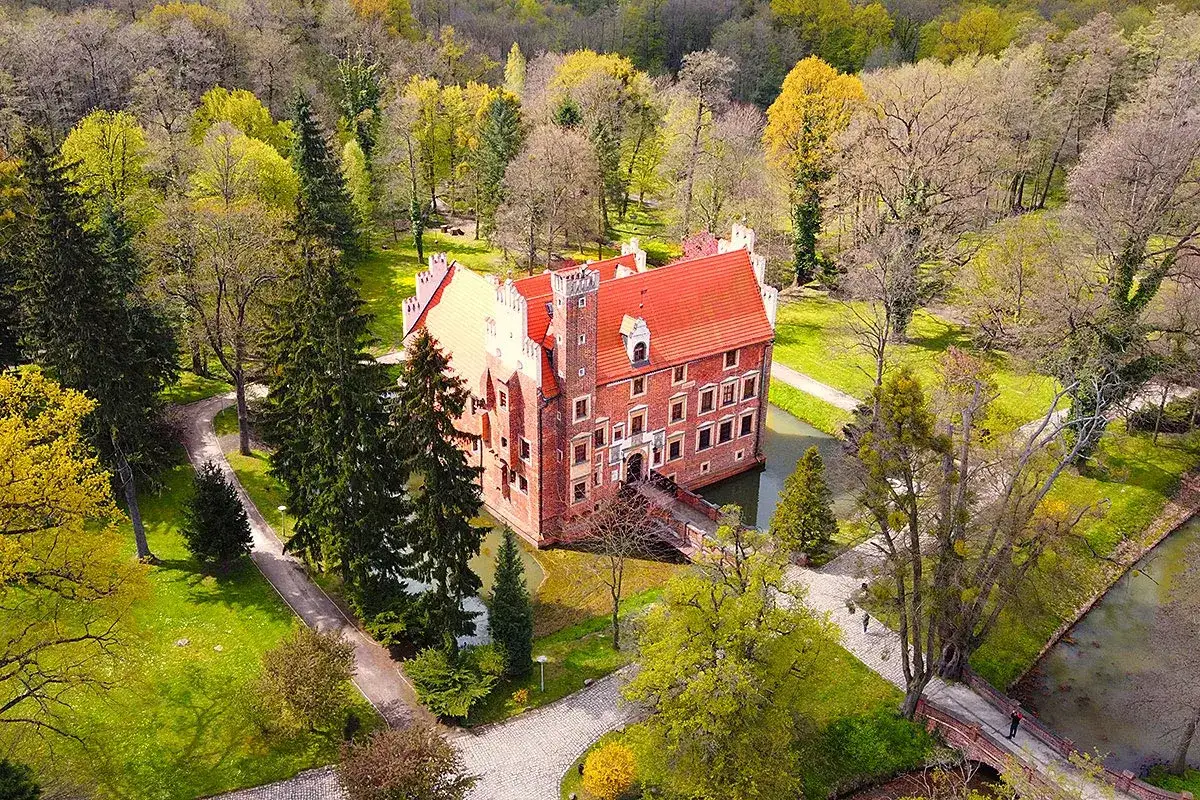 Zamek na wodzie w Wojnowicach otoczony wiosennym parkiem. Czerwona cegła i pomarańczowy dach lśnią w słońcu.