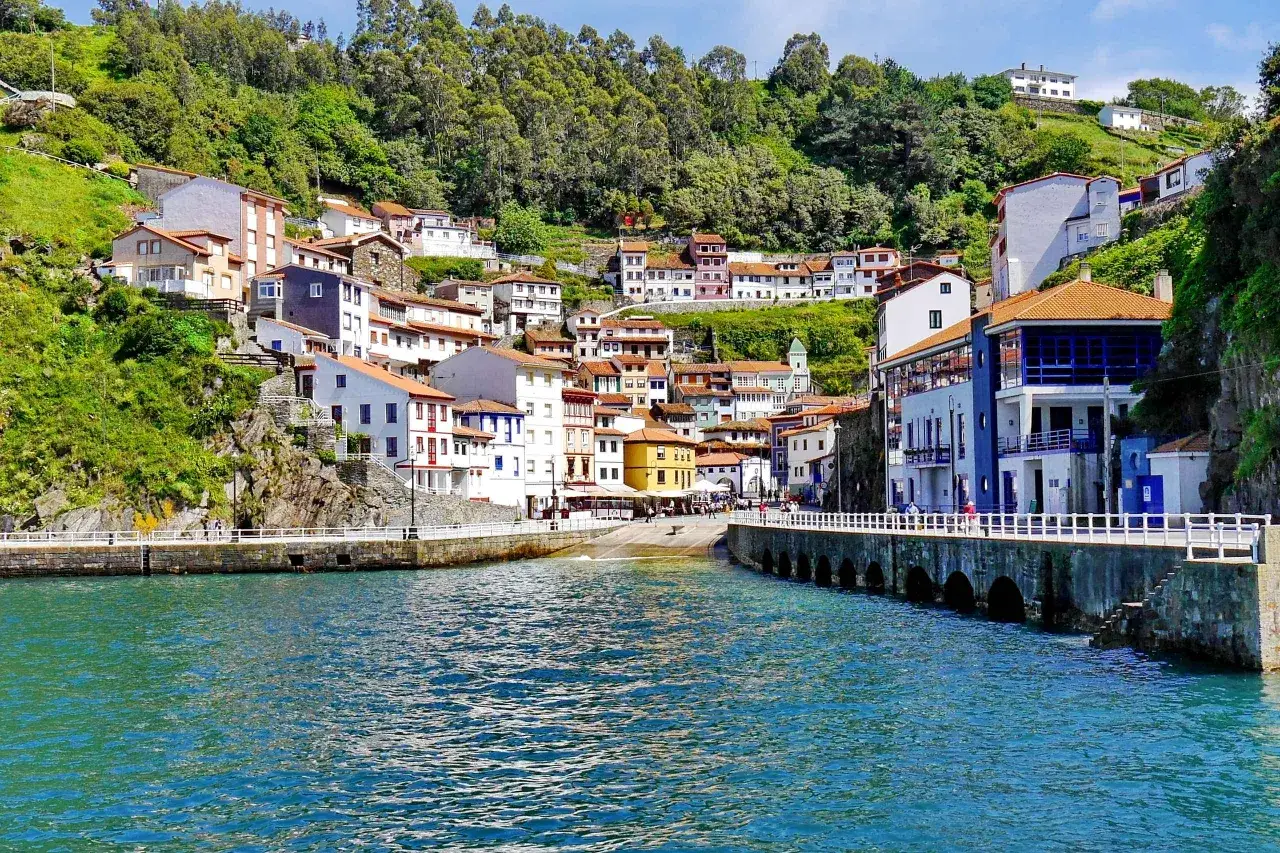 Bunte H&auml;user schmiegen sich an einen gr&uuml;nen Hang in Cudillero, Spanien. Das t&uuml;rkisfarbene Wasser des Hafens spiegelt die malerische Kulisse wider.