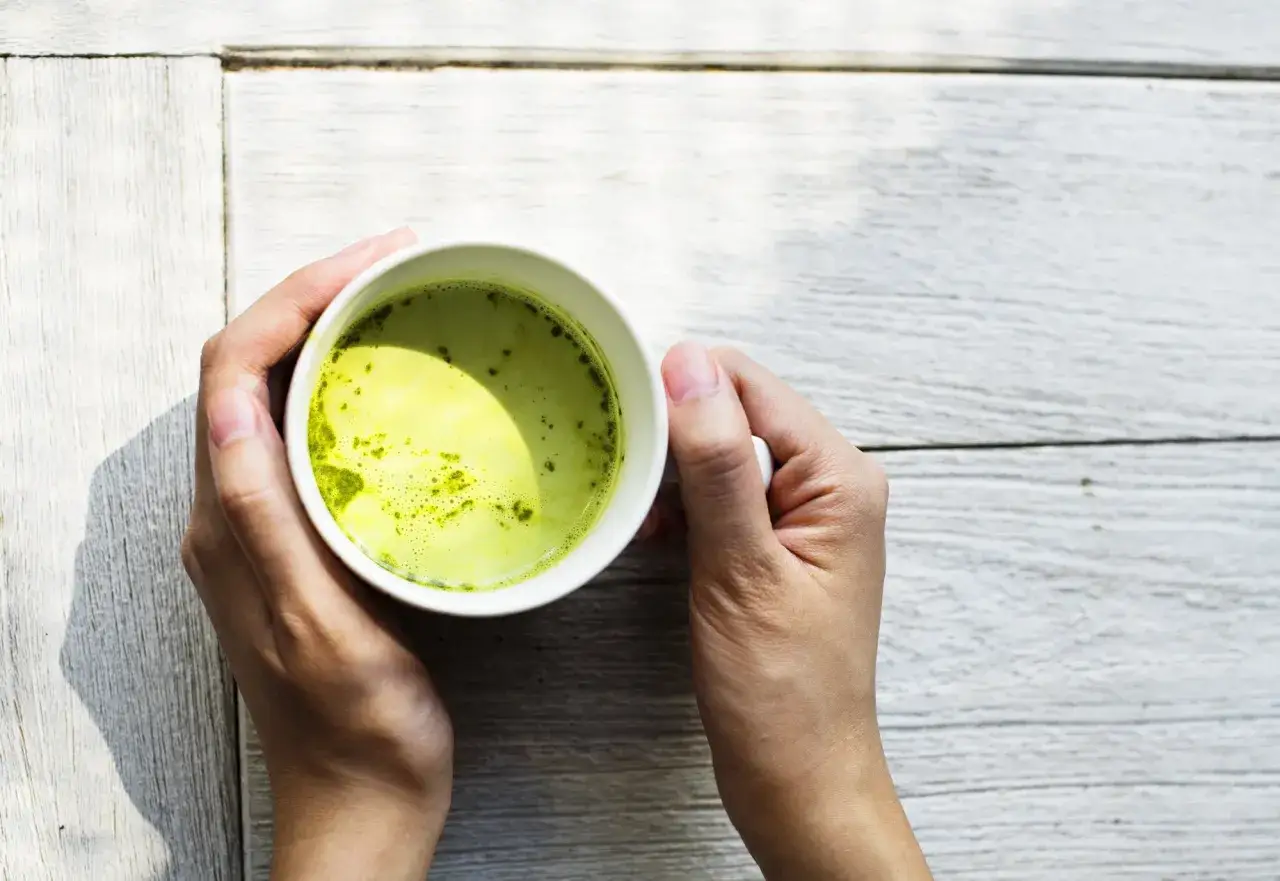 H&auml;nder h&aring;ller en kopp med gr&ouml;n matcha, en dryck rik p&aring; matcha koffein, p&aring; ett vitt tr&auml;bord.