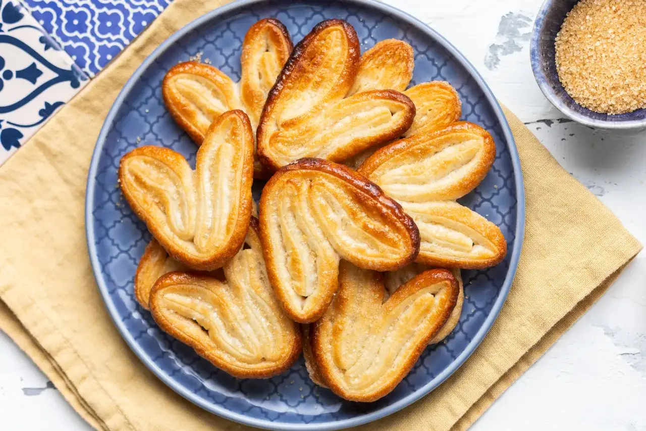 Un piatto blu pieno di dolci con pasta sfoglia dorati e croccanti, spolverati di zucchero.