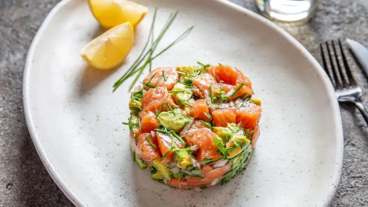 Un délicieux tartare de saumon classique, frais et coloré, servi avec des quartiers de citron et de la ciboulette.