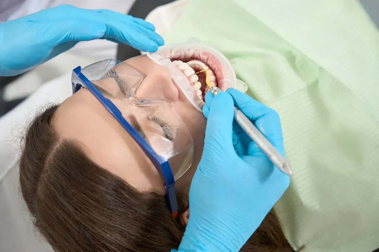 Dentysta wykonuje zabieg higienizacji jamy ustnej. Pacjentka ma założony koferdam i okulary ochronne.