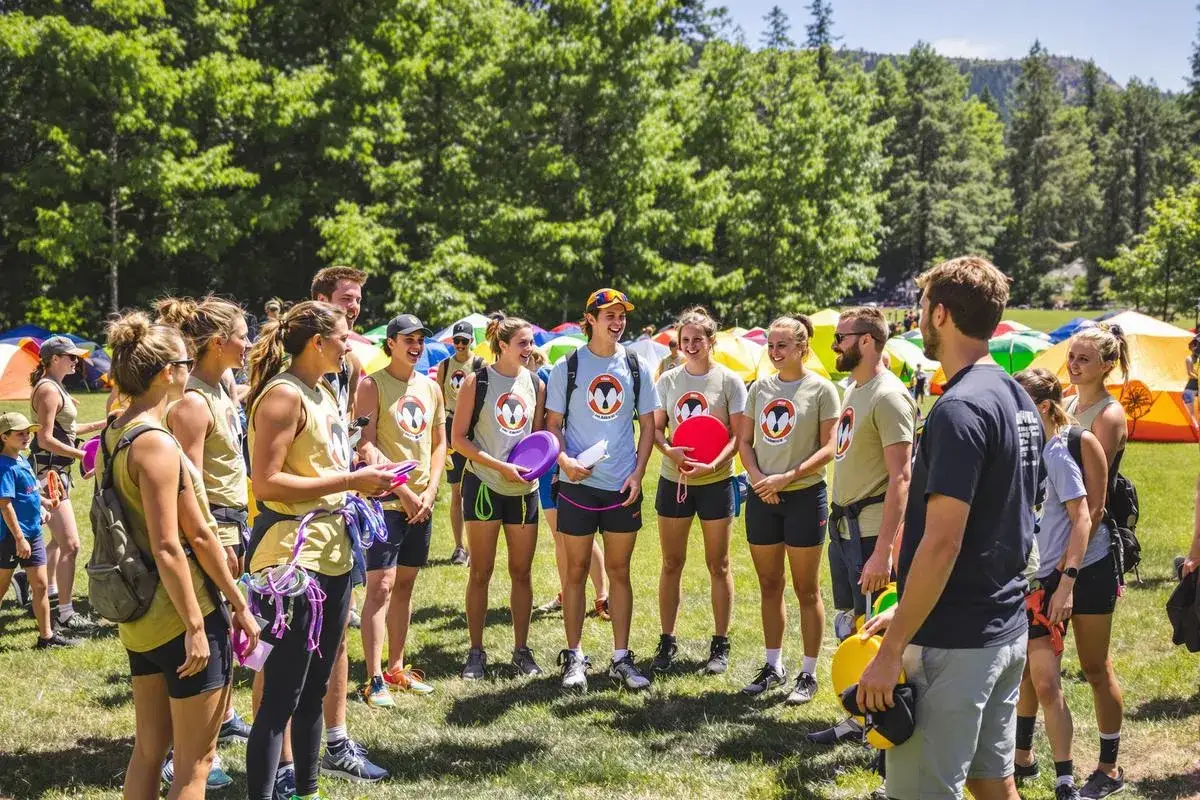 Un groupe de jeunes gens souriants, portant des t-shirts identiques, participe à un stage pratique en plein air. Ils tiennent des frisbees et du matériel d'escalade.