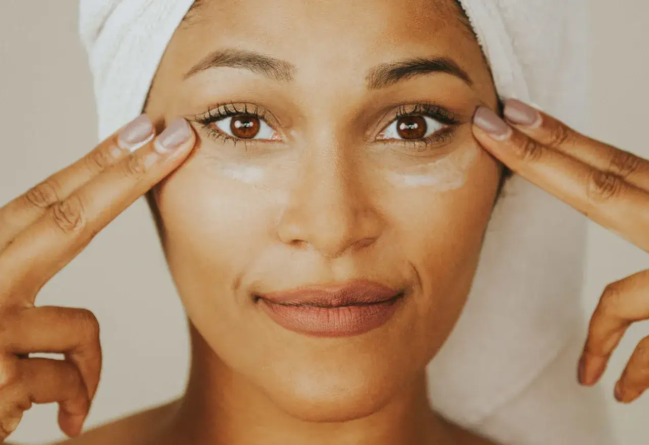 Une femme applique une crème contour des yeux sur tout le visage, avec une serviette sur la tête.