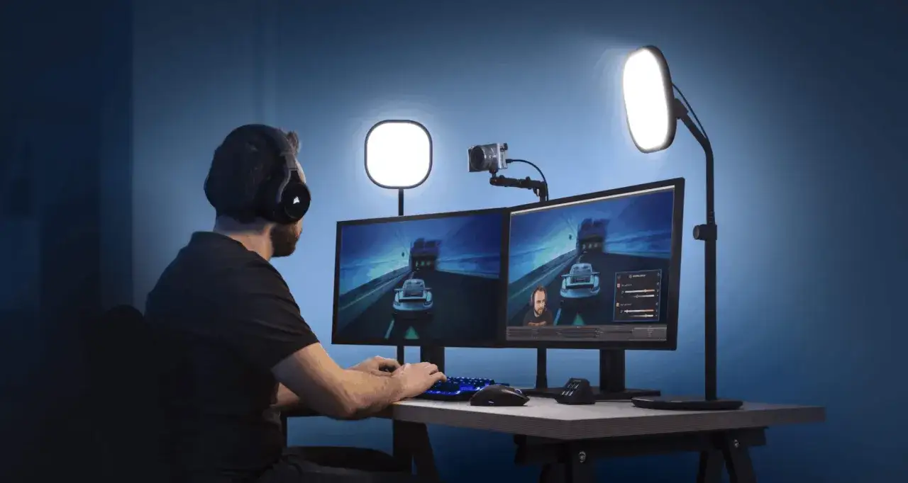 Person med h&ouml;rlurar spelar datorspel, med en kamera p&aring; datorn och tv&aring; sk&auml;rmar som visar spelet.