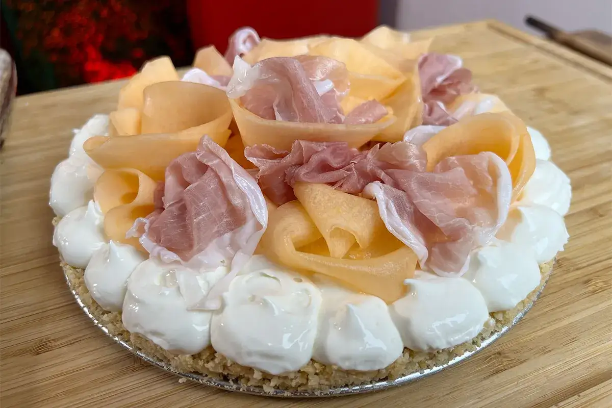Torta salata con base croccante, ricoperta di panna montata e decorata con fette di melone arrotolate e prosciutto crudo. Un'esplosione di sapori per un antipasto estivo.