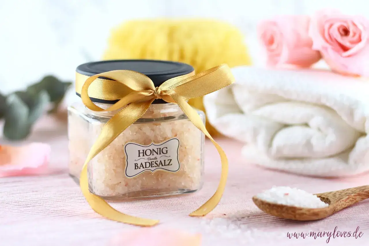 Ein Glas mit selbstgemachtem Honig-Vanille-Badesalz, verziert mit einem goldenen Band. Daneben liegen eine Holzschaufel mit Salz, eine weiche B&uuml;rste und gefaltete Handt&uuml;cher.