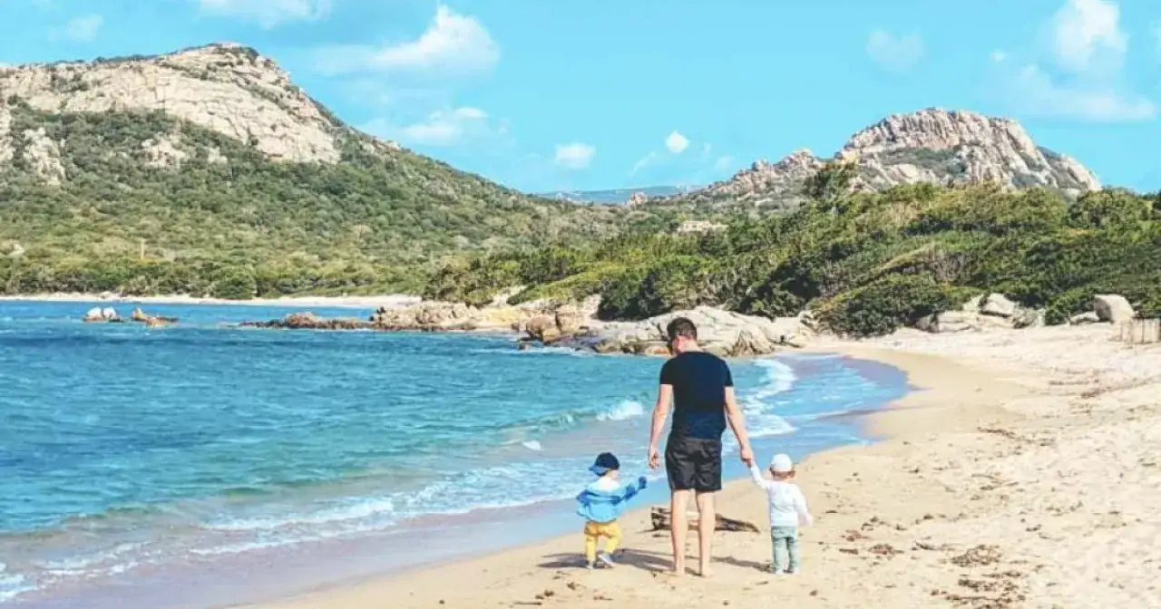 Un père et ses deux enfants marchent sur une plage de sable fin, face à la mer bleue et des montagnes verdoyantes. Idéal pour savoir où partir en famille en juillet.